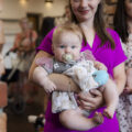 Na zdjęciu widzimy mamę trzymającą niemowlę w ramionach. W tle inni rodzice.