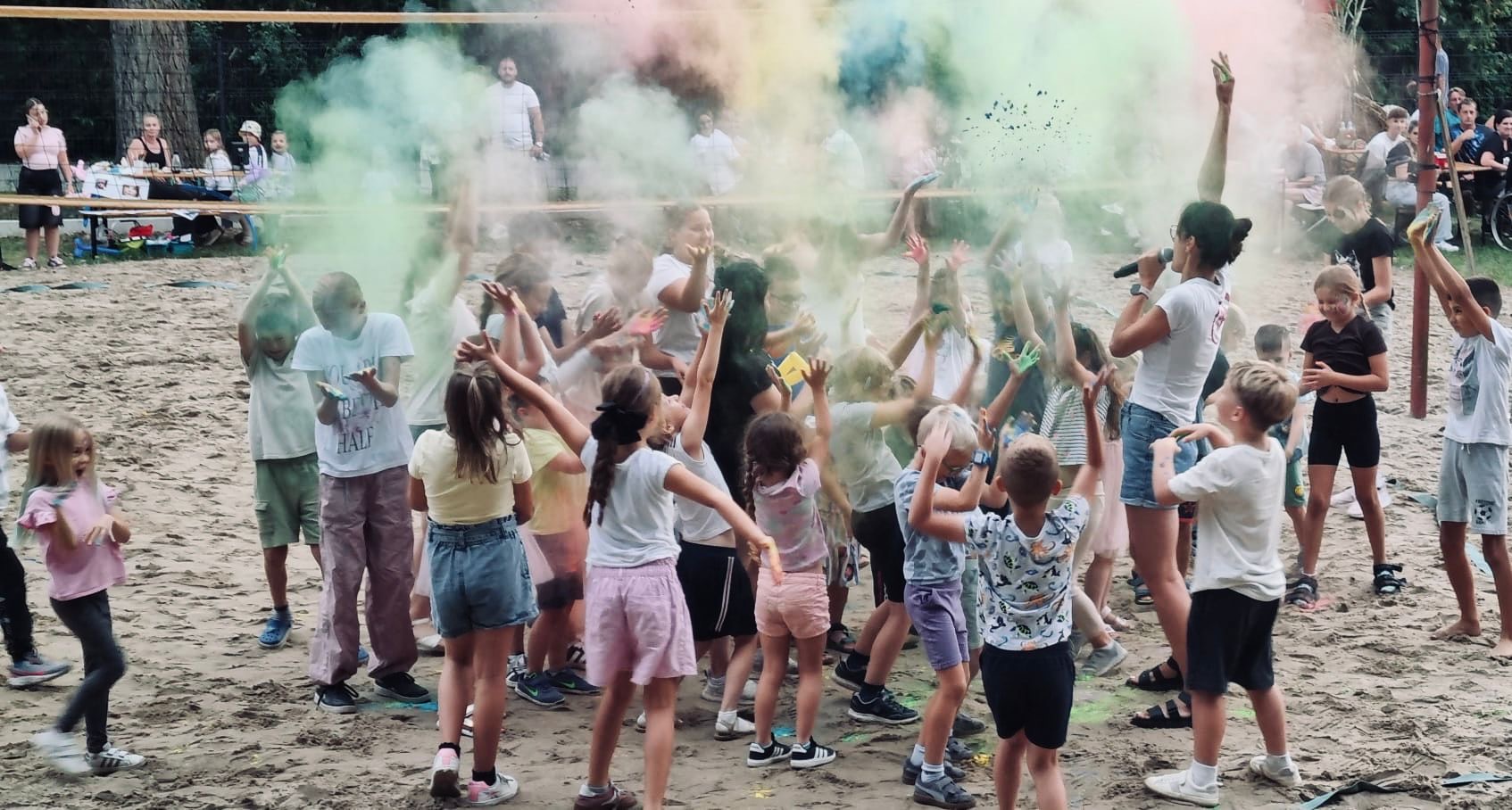 Zdjęcie przedstawia dzieci podczas festiwalu kolorów. Na piaskowym boisku bawi się duża grupa dzieci. W powietrzu unosi się chmura kolorowego proszku. Dzieci klaszczą, skaczą, rzucają kolory w górę i śmieją się. Z boku stoi kobieta z mikrofonem, która prowadzi zabawę.