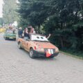 Na zdjęciu auto w korowodzie, które udekorowane zostało tak, że wygląda jak postać z bajki „Auta” – Złomek i taki też ma napis na drzwiach. Ma namalowane wielkie oczy na szybie i czerwony język wystający z przodu maski. Z wnętrza auta machają wesoło uczestnicy wydarzenia. Za nim widać inne przystrojone pojazdy.