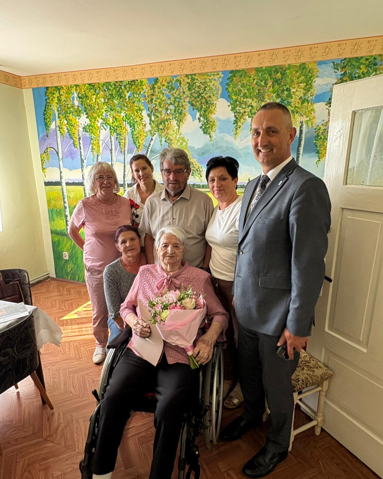 Na fotografii wójt, jubilatka oraz jej rodzina. Jubilatka trzyma bukiet kwiatów i list gratulacyjny.