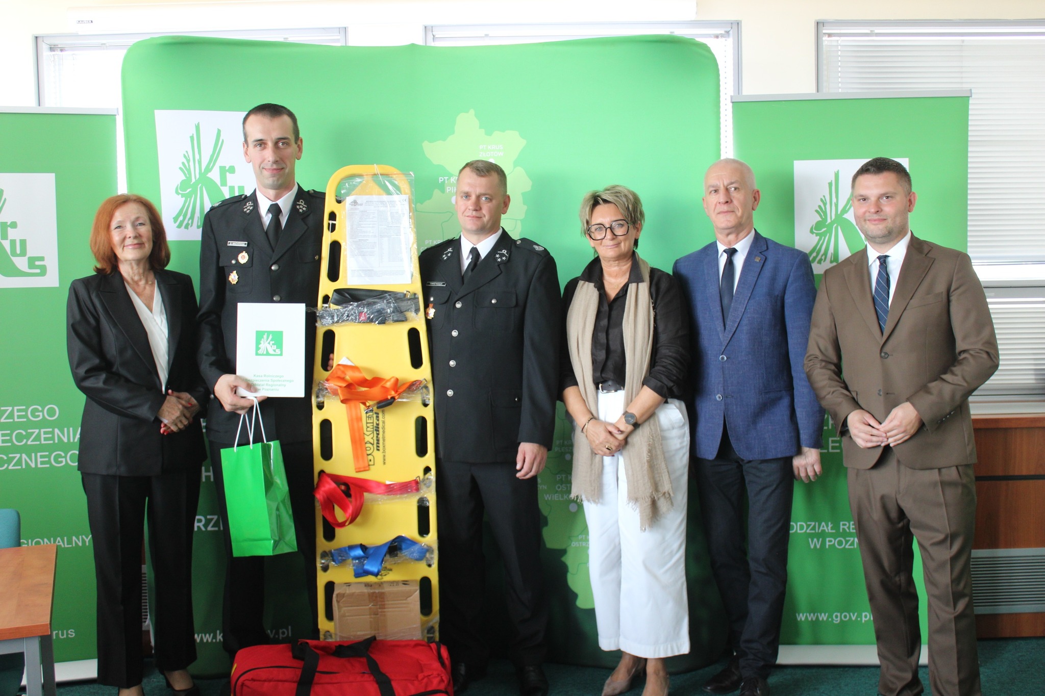 Na fotografii osoby ze straży pożarnej oraz przedstawiciele KRUS pozują z nowoczesnym zestawem ratownictwa przedlekarskiego - torbą medyczną i deską ortopedyczną.