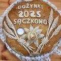 Na zdjęciu widoczny jest tradycyjny chleb dożynkowy, udekorowany misternymi zdobieniami z ciasta – kłosami zbóż, liśćmi oraz kwiatami. Na wierzchu umieszczony jest napis: „Dożynki 2025 Sączkowo”. Chleb spoczywa na koronkowej serwetce.