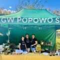 Na fotografii trzy kobiety z KGW Popowo Stare pozują przy stanowisku z przygotowanym przez siebie jedzeniem. Nad nimi rozpościera się duży zielony namiot z napisem "KGW Popowo Stare".