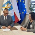 Na zdjęciu wójt podczas podpisywania umowy. Koło niego siedzi skarbnik gminy. W tle flagi: Polski, UE i flaga z herbem gminy Przemęt.