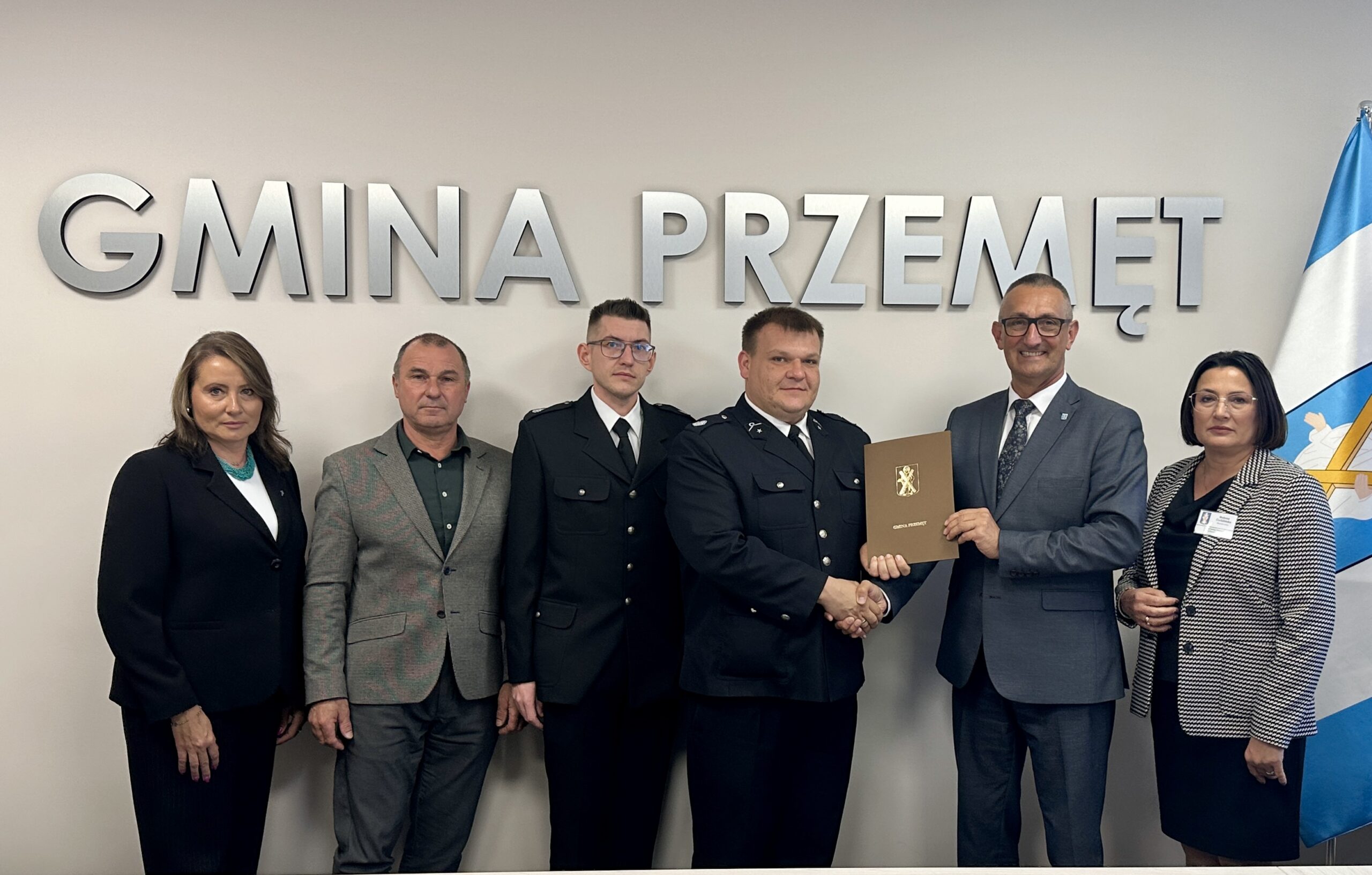 Wspólne zdjęcie, na którym znajdują się wójt Janusz Frąckowiak, skarbnik gminy Bożena Ćwiklińska, prezes OSP Bucz Robert Pawłowski, skarbnik jednostki Sławomir Krystek, a także radni Rady Gminy Przemęt: Anna Barczyńska oraz Jerzy Klecha. Wójt oraz prezes OSP podają sobie dłoń oraz wspólnie trzymają teczkę, w której znajduje się umowa. W tle srebrny napis: "Gmina Przemęt".