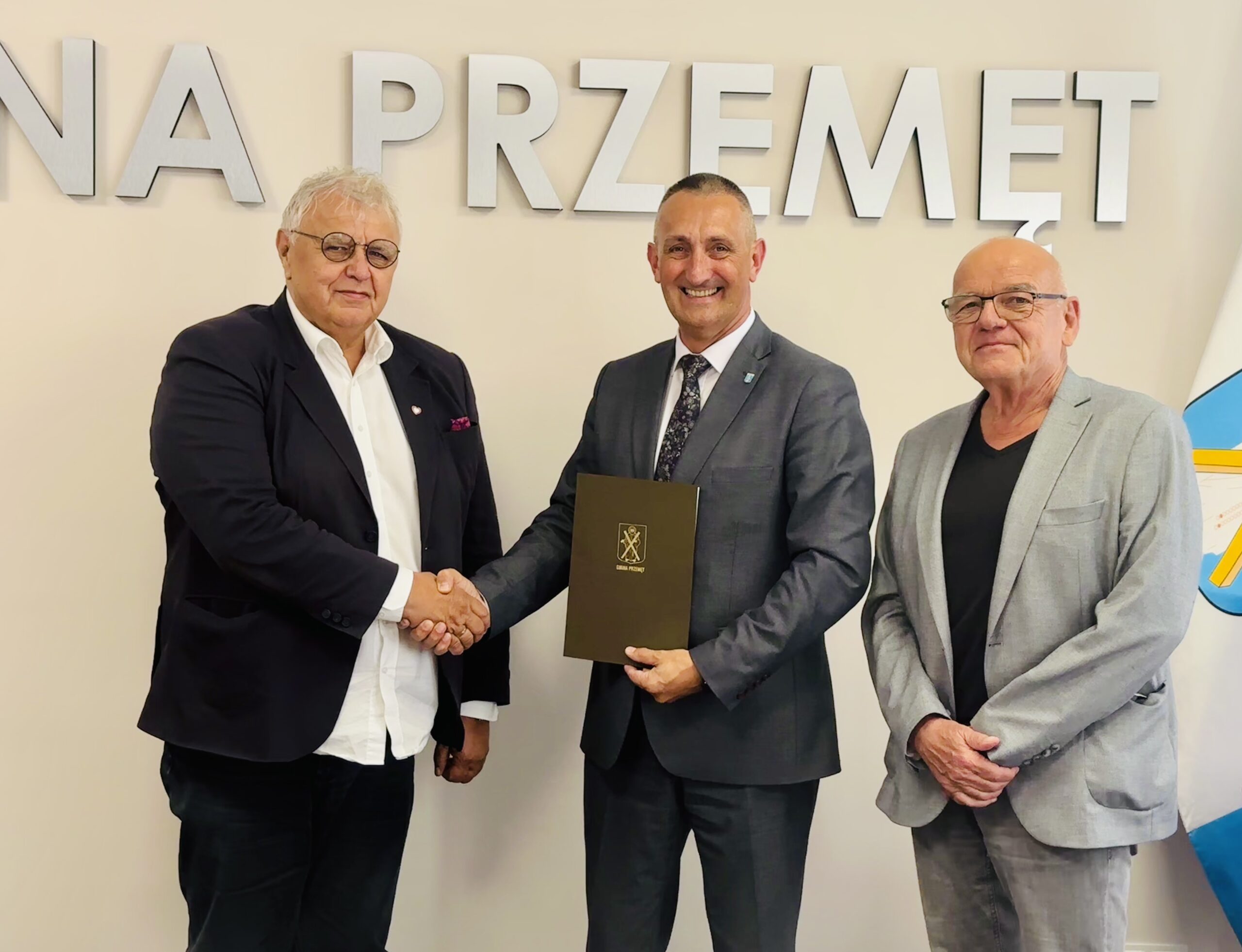 Na fotografii wójt Janusz Frąckowiak oraz dyrektor Karol Mońko. Wyżej wymienione osoby podają sobie dłoń. Wójt trzyma teczkę, w której znajduje się umowa. Do zdjęcia pozuje także dr Stanisław Wyrzykowski. W tle srebrny napis: "Gmina Przemęt".