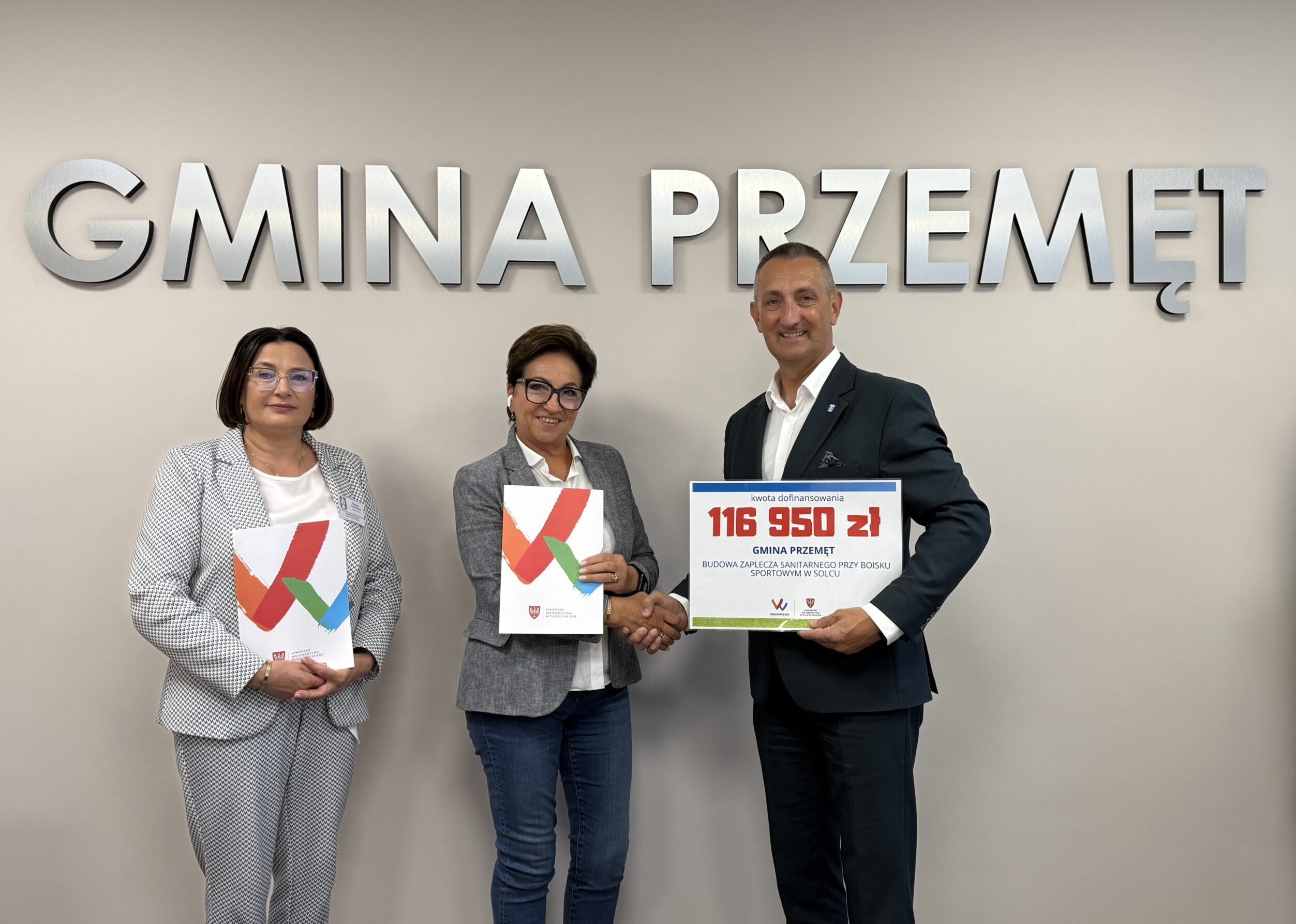Na fotografii wójt, skarbnik oraz pani Barbara Mroczkowska pozują na tle srebrnego napisu „Gmina Przemęt”. Wójt i pani Barbara podają sobie dłonie. Wójt trzyma tabliczkę z widniejącą na niej kwotą 116 950 zł, natomiast pani Barbara trzyma teczkę z umową. Jeden egzemplarz umowy znajduje się także w teczce trzymanej przez panią skarbnik.