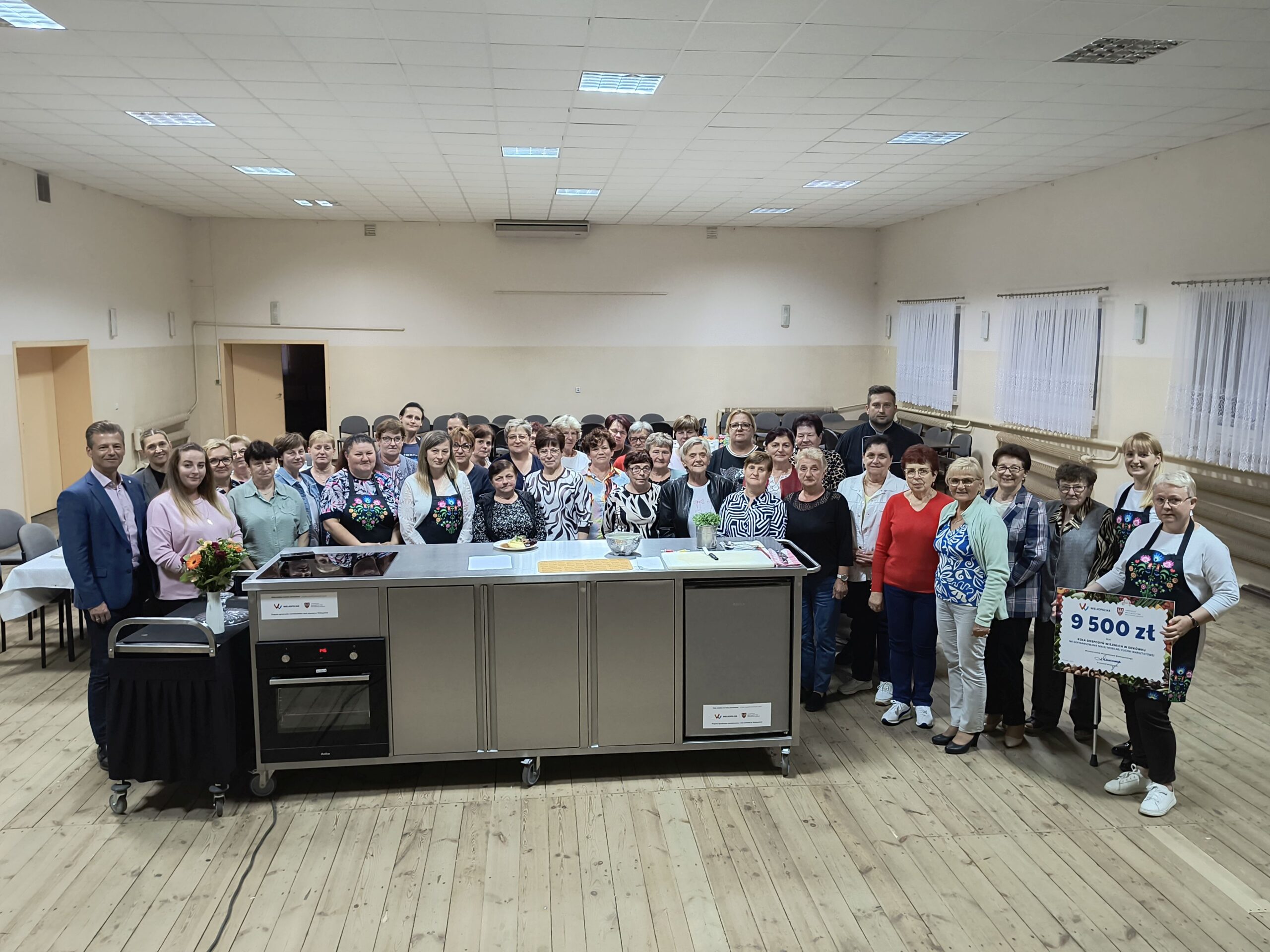 Grupowe zdjęcie, na którym widoczni są: uczestnicy warsztatów, kucharz, panie z KGW, wicewójt Waldemar Kalitka oraz Elwira Michalewicz. Wszyscy pozują przy Mobilnej Kuchni Warsztatowej.