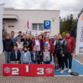 Fotografia przedstawia uczestników po zakończonym biegu, pozujących z medalami i pucharem. Wśród zgromadzonych znajdują się również osoby, które nie brały udziału w biegu. Przed grupą ustawione jest sportowe podium, a obok stoją windery informujące o honorowym patronacie wójta gminy.