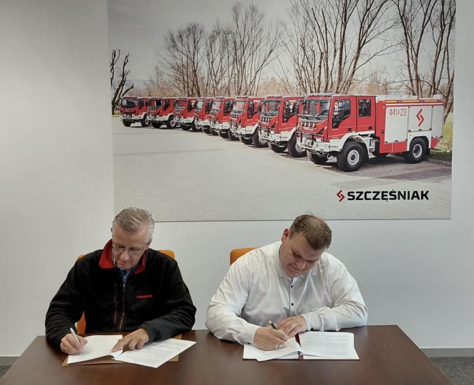 Na zdjęciu prezes jednostki OSP Robert Pawłowski oraz właściciel firmy "SZCZĘŚNIAK". Wyżej wymienieni podpisują umowę. W tle obraz z wozami strażackimi.