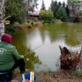 Na fotografii widoczne są osoby biorące udział w zawodach wędkarskich, siedzące przy swoich stanowiskach nad stawem.