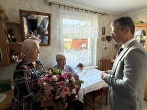 Na fotografii zastępca wójta wręcza bukiet kwiatów i składa życzenia pani Irenie. W tle na krześle siedzi pan Władysław.