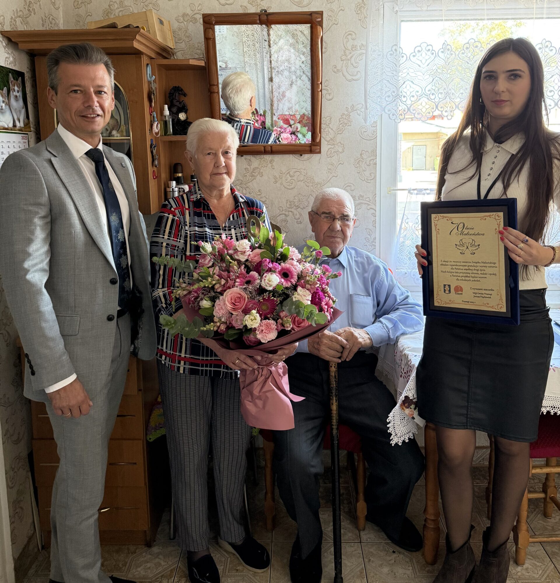 Na fotografii jubilaci wraz z zastępcą wójta i panią Natalią, która pracuje w urzędzie stanu cywilnego w gminie. Jubilatka trzyma bukiet kwiatów. Pani Natalia trzyma grawerton z życzeniami z okazji 70-lecia ślubu.