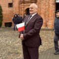Na zdjęciu widoczny jest mężczyzna stojący przed kościołem i trzymający małą flagę Polski na patyczku.