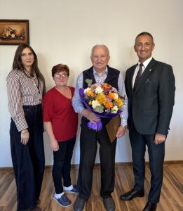 Wspólne zdjęcie, na którym znajdują się wójt, pracownik Urzędu Stanu Cywilnego w Przemęcie oraz jubilat z członkiem swojej rodziny. Jubilat pozuje z bukietem kwiatów i listem gratulacyjnym.