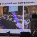 Na zdjęciu widoczna kobieta z KGW Siekówko, która przemawia na scenie. Opowiada ona o działalności KGW. Na projektorze wyświetlane są zdjęcia z jubileuszu 65-lecia KGW Siekówko.