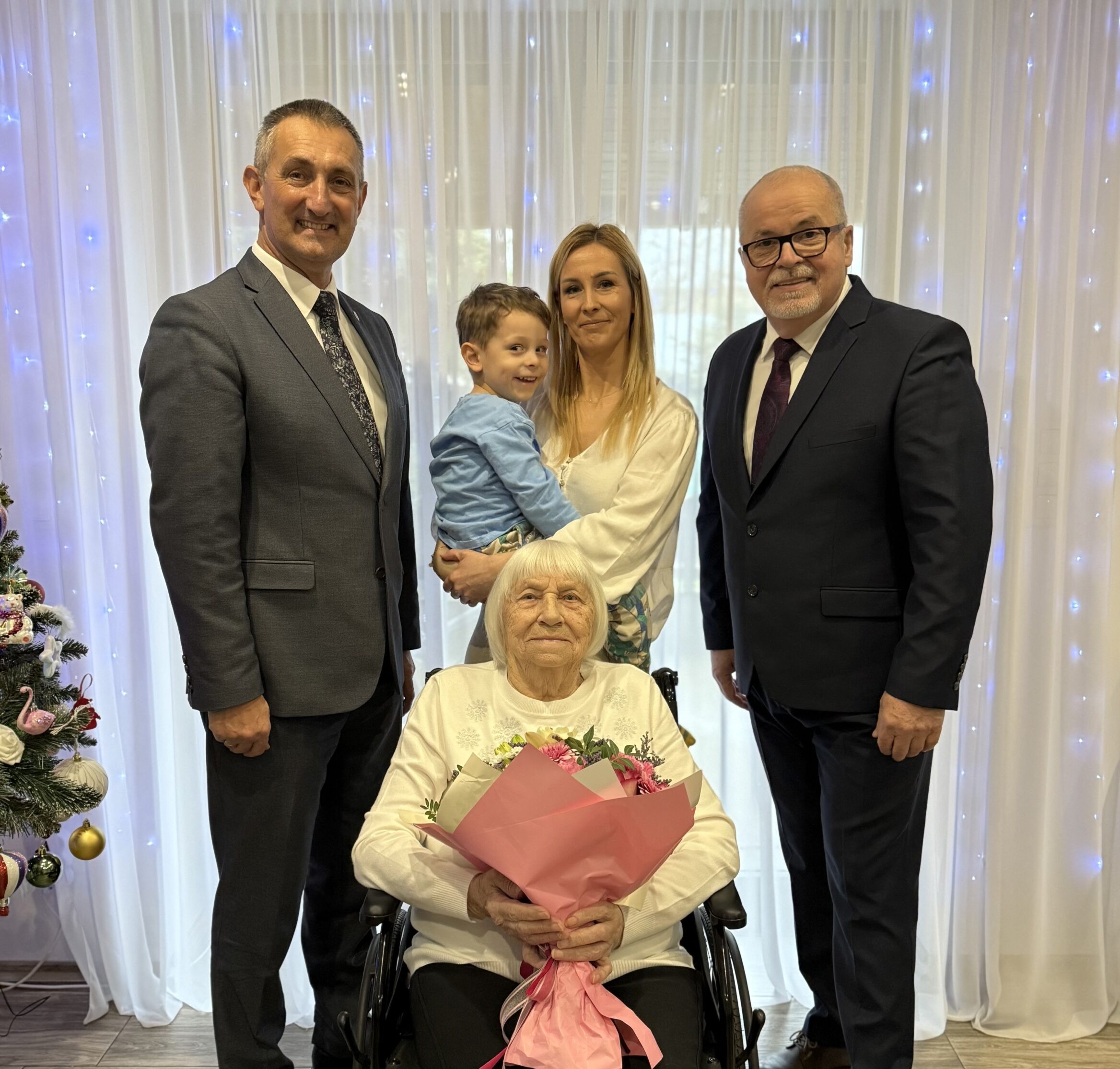 Grupowe zdjęcie, na którym znajdują się wójt, kierownik USC oraz jubilatka wraz z bliskimi. Jubilatka pozuje z bukietem kwiatów.
